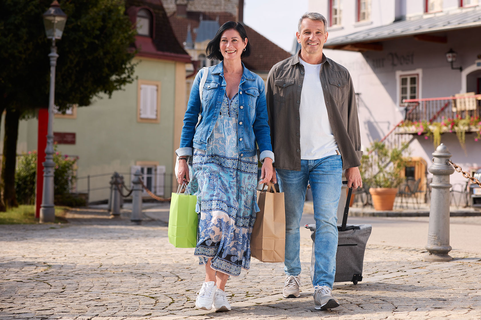 Mann und Frau gehen mit Einkaufstaschen und -trolley über einen öffentlichen Platz.