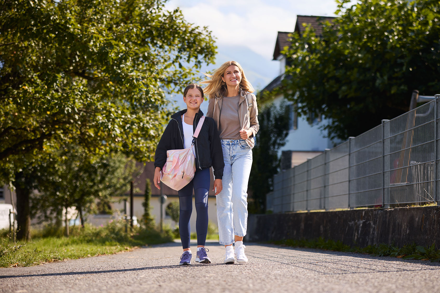 Eine Frau geht mit ihrer Tochter auf einem Fußgängerweg. Das Mädchen trägt eine Sporttasche