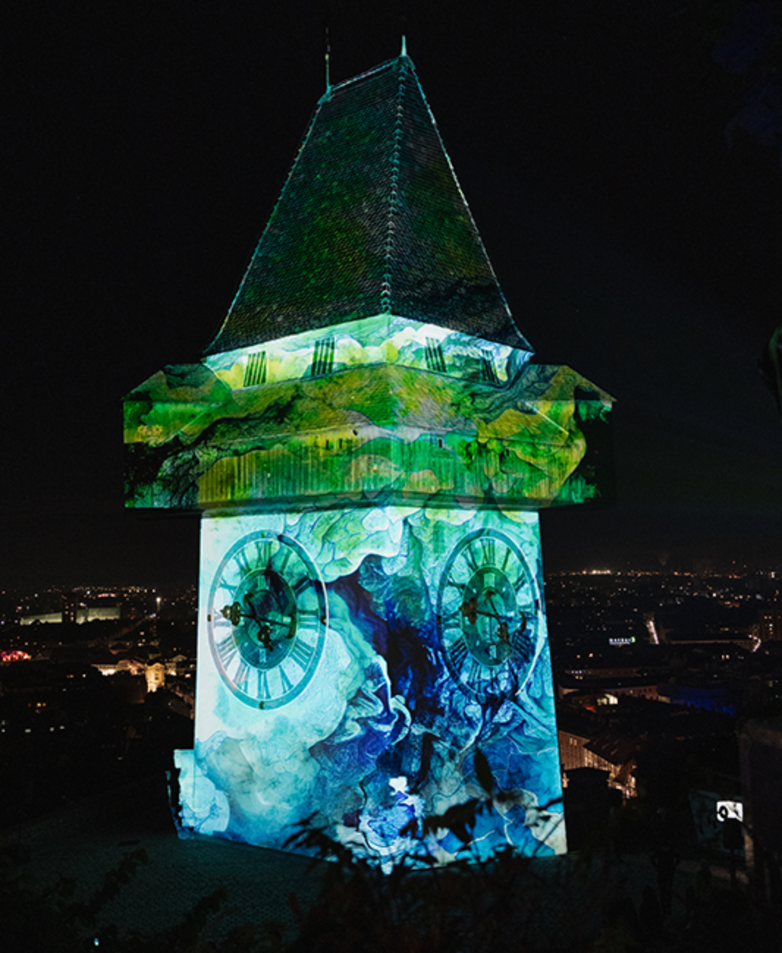 Der Grazer Uhrturm bei Nacht, beleuchtet mit farbigen Bildern.