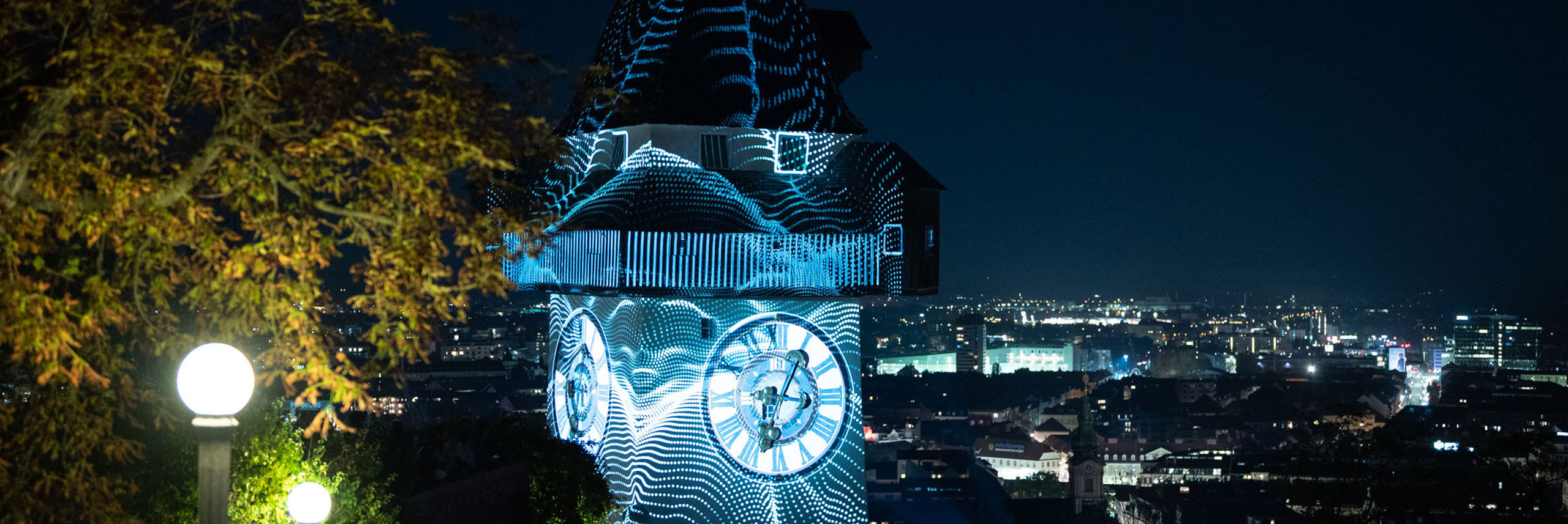 Ein Uhrturm bei Nacht, über den eine Lichtprojektion geblendet wird.