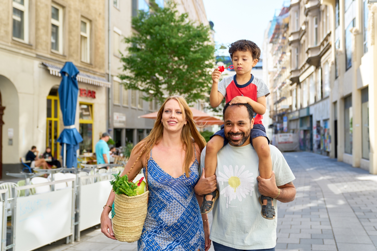 Frau mit Einkaufstasche und Mann mit Kind auf den Schultern spazieren durch die Innenstadt in Wien