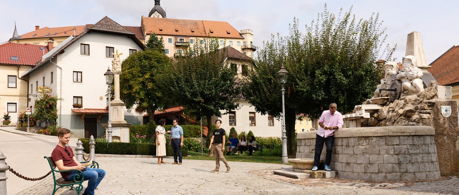 Ein Platz mit gepflastertem Stein. Rechts ein Brunnen auf dem ein Mann lehnt und aufs Handy schaut, links ein Mann auf einer Bank, der ein Buch liest. Im Hintergrund gehende Menschen, die miteinander reden.