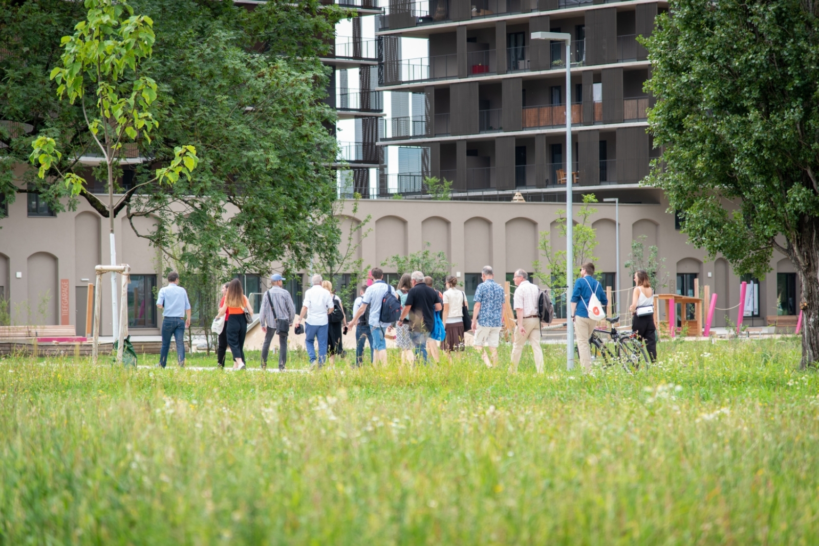 Viele Personen in der Ferne, die zu Fuß gehen, vorne viel grüne Wiese, im Hintergrund eine Siedlung