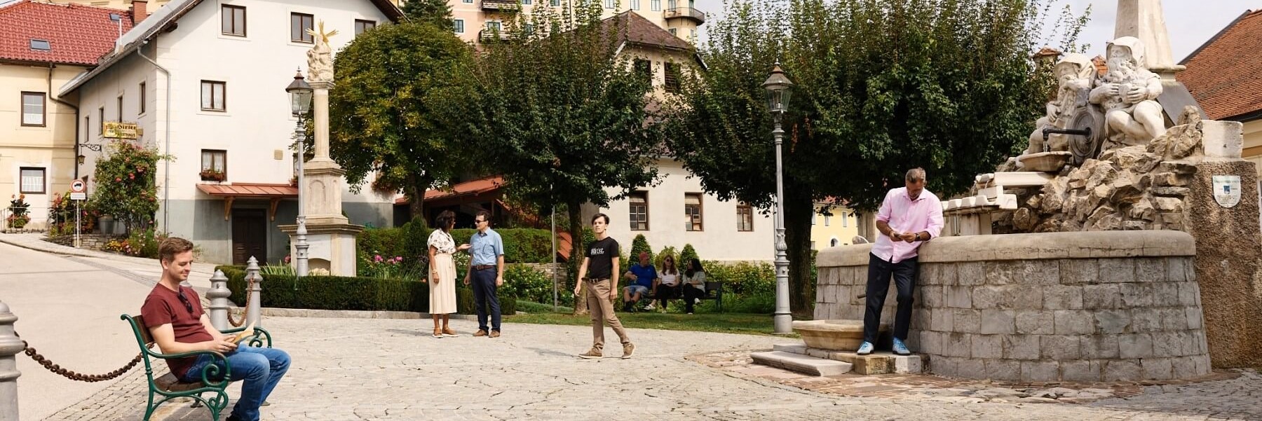 Viele Personen auf einem Platz, links liest jemand, rechts schaut jemand aufs Handy und lehnt an einem großen Zierbrunnen, hintn reden zwei Personen