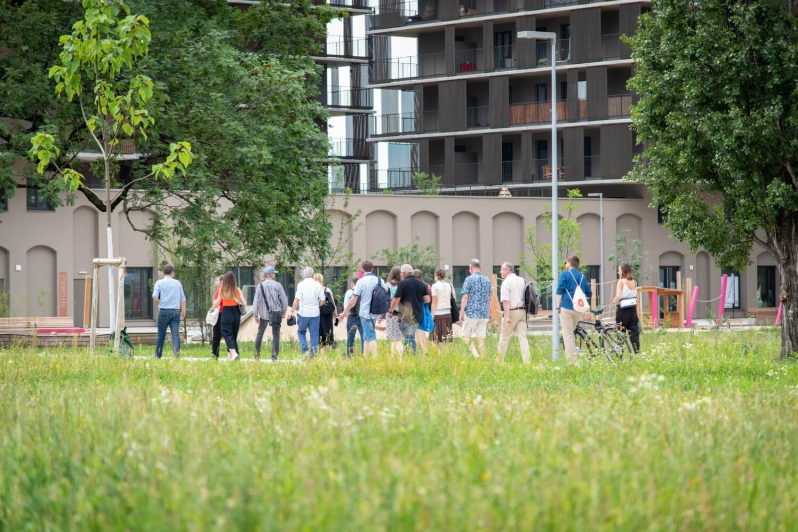 Viele Personen in der Ferne, die zu Fuß gehen, vorne viel grüne Wiese, im Hintergrund eine Siedlung