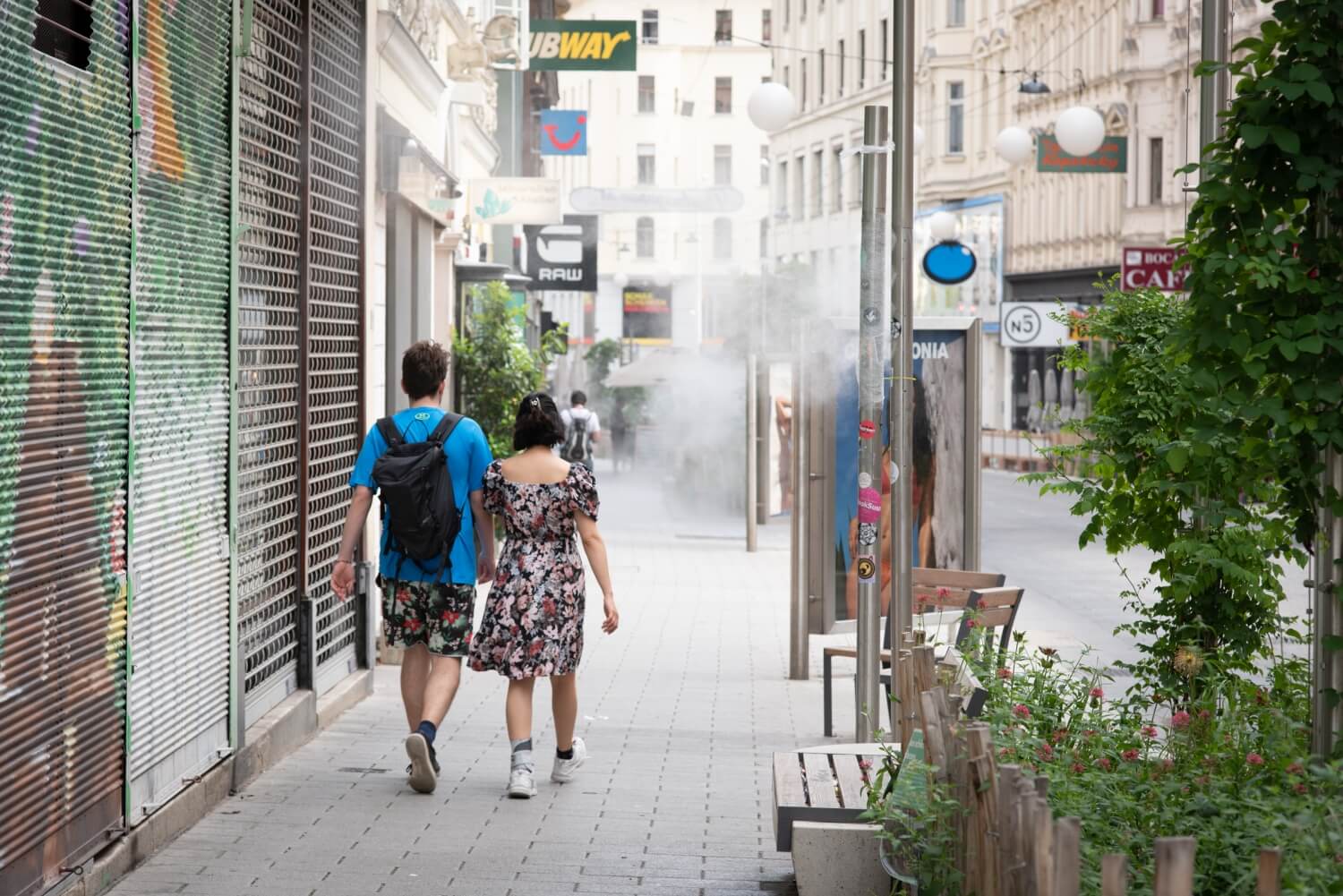 Ein paar geht entlang einer Fußgängerzone - links ein mit Gitter geschlossenes Geschäft, vorne eine Nebeldusche, rechts Begrünung und Sitzmöglichkeiten.
