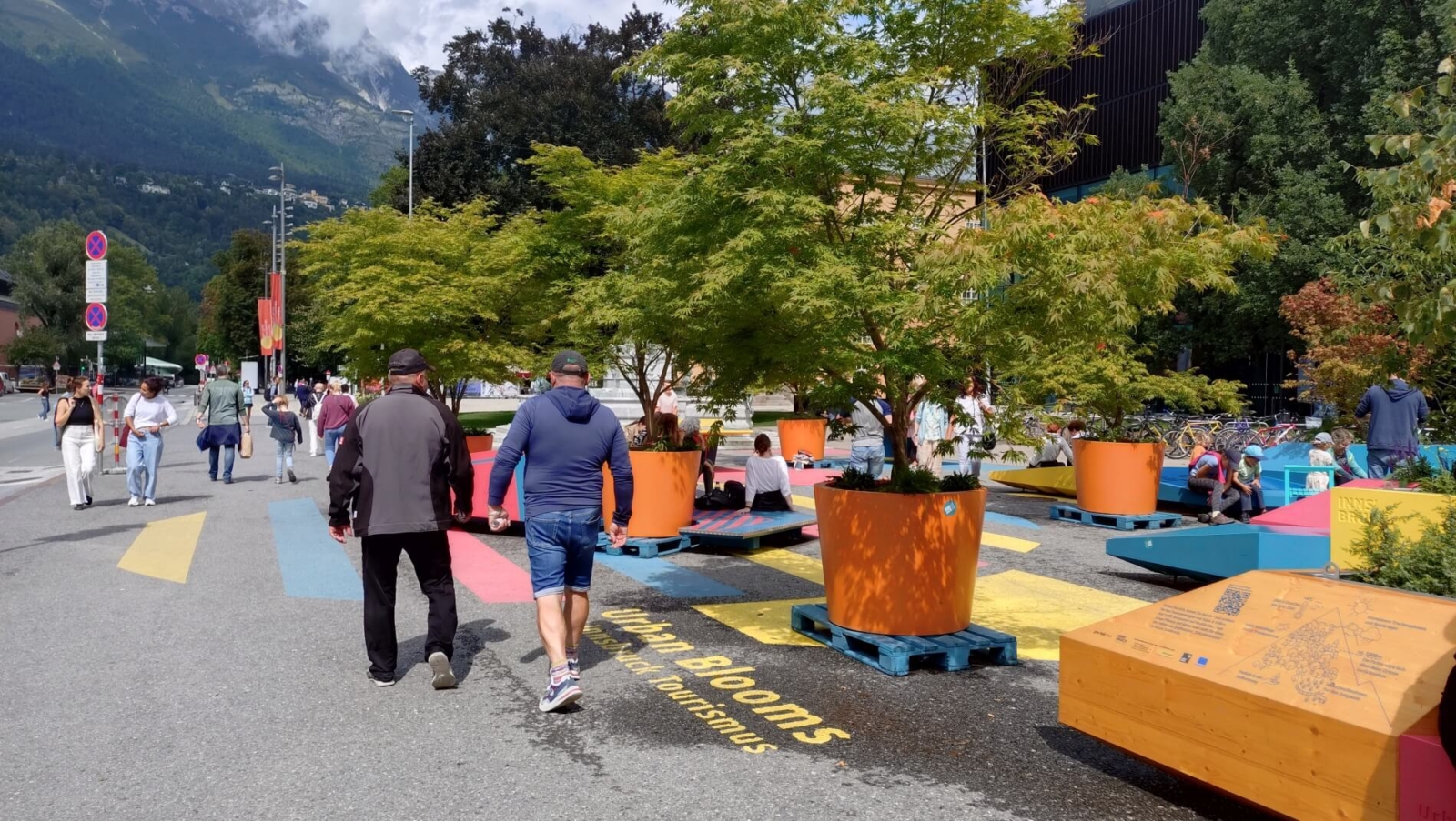 bunte Sitzmöbel auf einem Platz, dazwischen große Stauden in Blumentöpfen. Menschen liegen darauf und gehen über den Platz.
