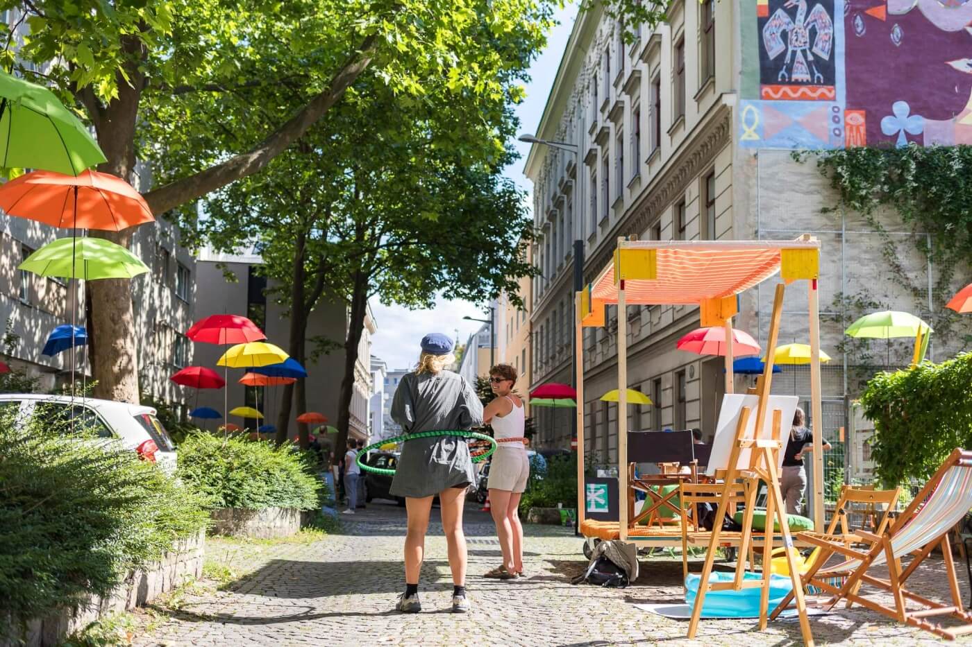 Eine blühende Wohnstraße mit Klappstühlen, bunten Schirmen und zwei Menschen, die Hoola-Hup-Reifen drehen