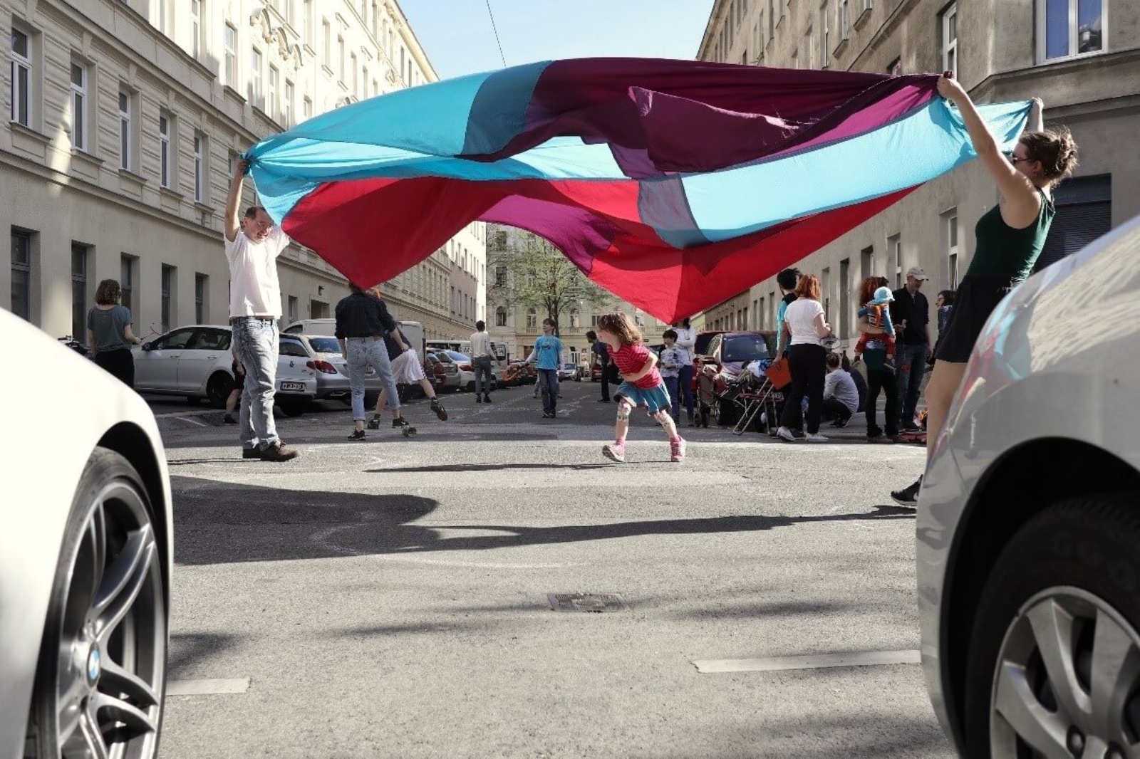 Durch zwei parkende Autos hindurch geschaut sieht man auf der Mitte der Straße zwei Erwachsene mit Schwungtuch in der Luft und darunter ein Kind durchlaufen.