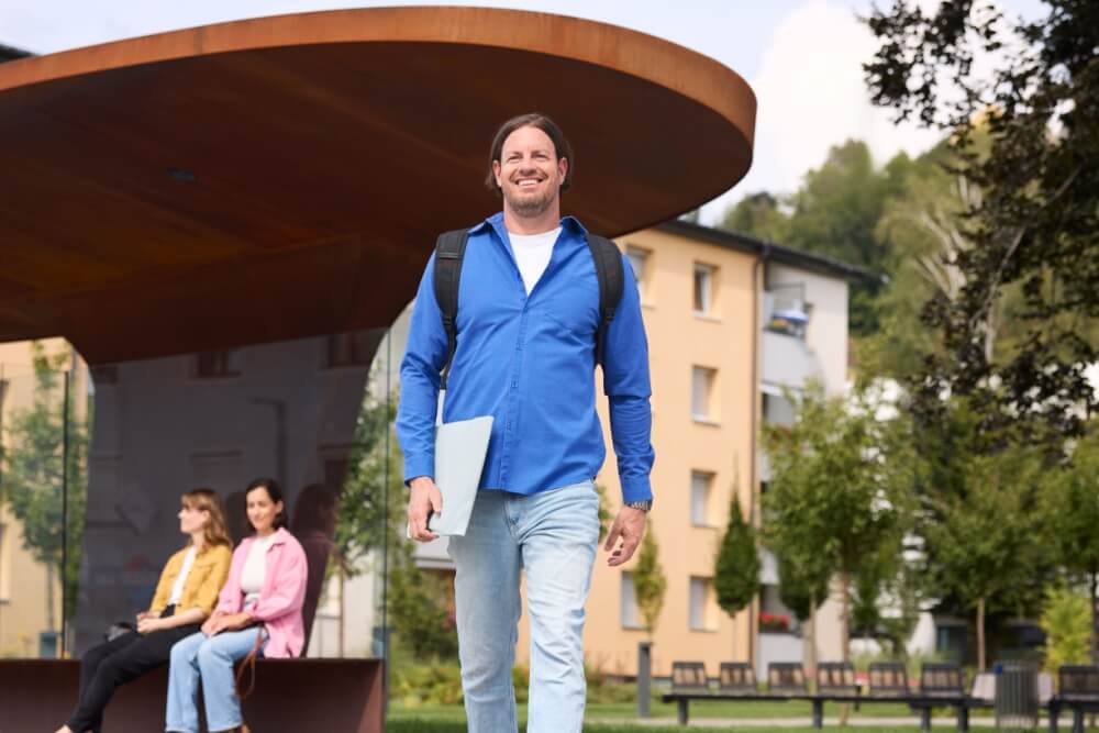 Mann mit Mappe in der Hand geht, im Hintergrund eine moderne Bushaltestelle mit kurvigem Flachdach. Darunter sitzen zwei Personen und warten.
