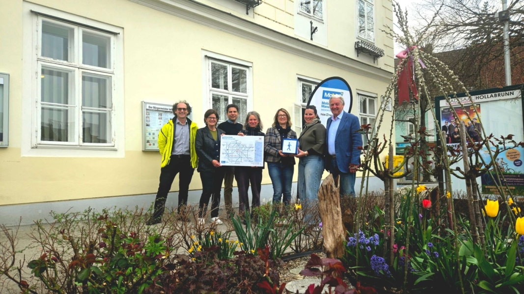 Mehrere Personen posieren vor einem Gebäude, im Vordergrund ein Blumenbeet. Sie halten ein Schild