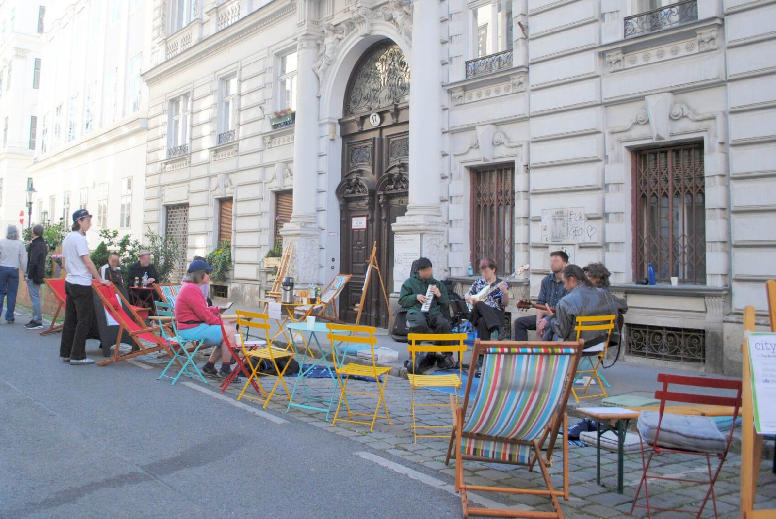 Mehrere Personen sitzen auf bunten Sesseln, welche auf einem Parkplatz stehen. Im Hintergrund ein typisches Wiener Haus mit Stuck.