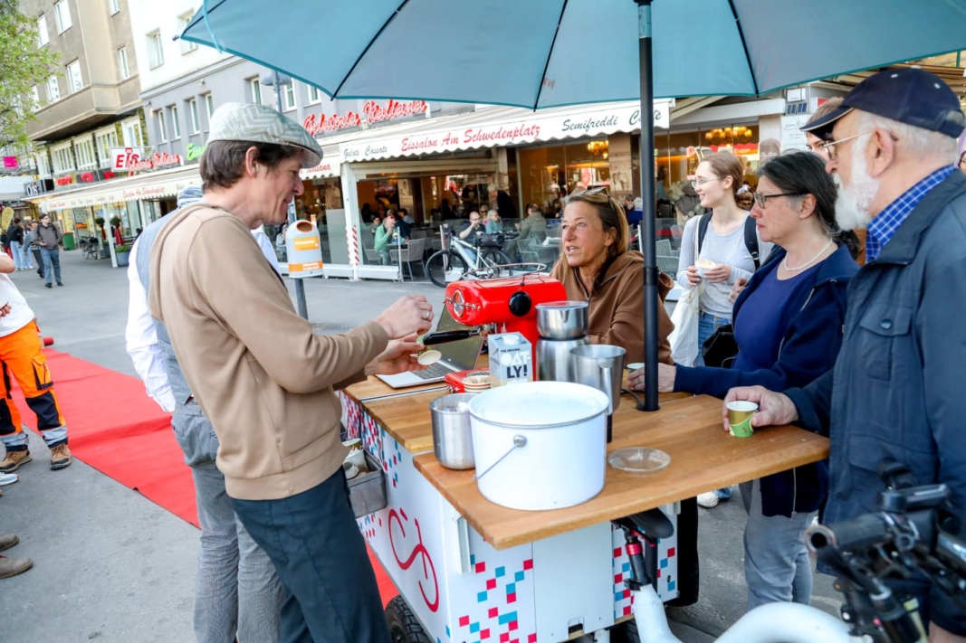 Ein kleiner Standtisch mit Schirm und einer Kaffemaschine, rundherum Menschen