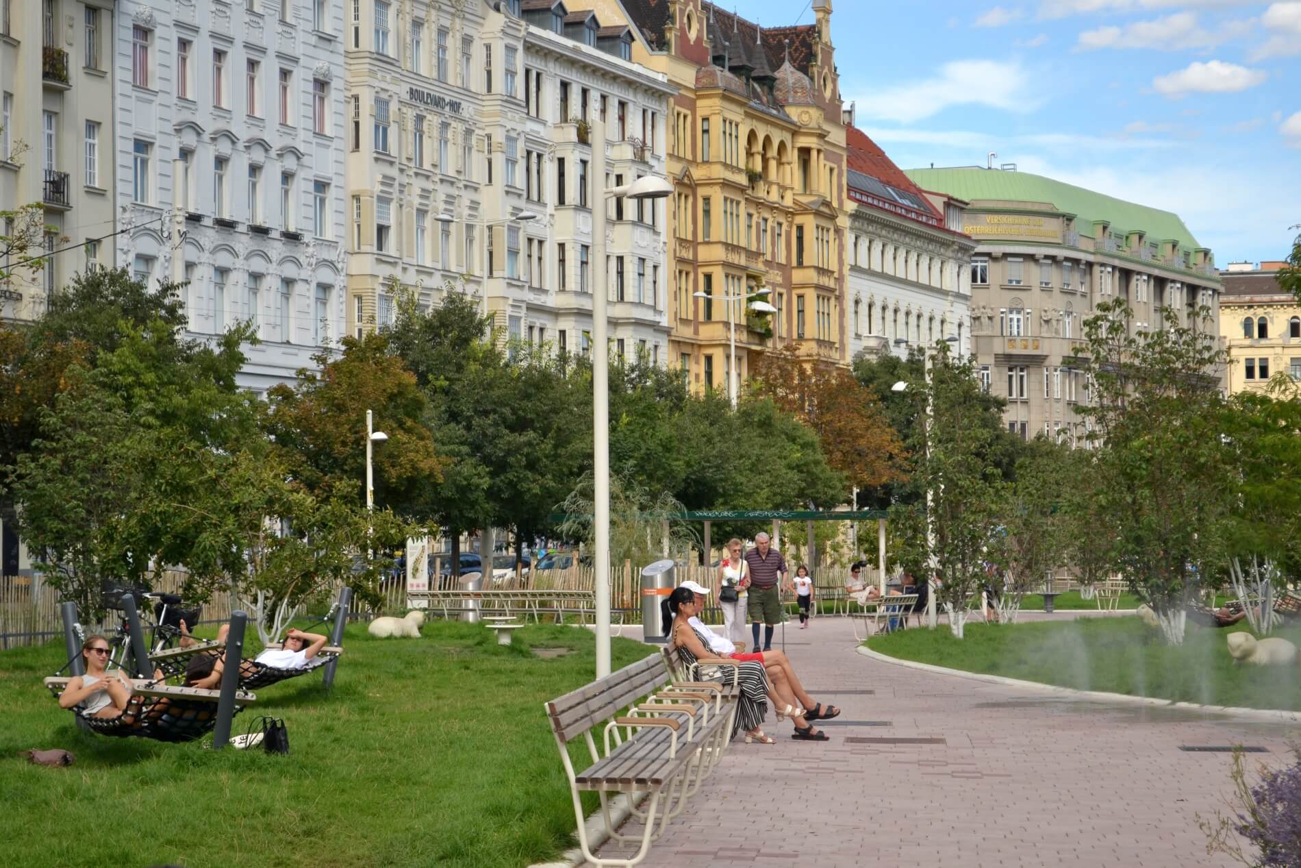 Wiener Naschpark mit Hängematten links in der Wiese, Bänke und Gehwegen mit Passanten und Nebelduschen. Im Hintergrund die Wienzeile Gebäude